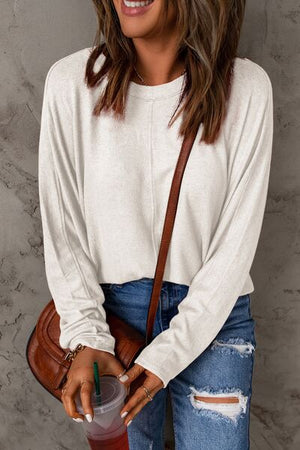 Smiling woman in a cream long sleeve T-shirt, holding a drink and wearing stylish jeans and a brown shoulder bag.