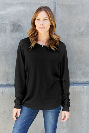 Model wearing a black round neck long sleeve t-shirt, paired with blue jeans, standing against a gray wall.