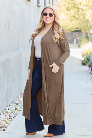 Stylish woman in a long brown cover-up with pockets, wearing sunglasses and flared jeans, walking outdoors.