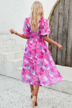 Back view of a woman in a smocked printed V-neck short sleeve dress in pink and purple hues, showcasing a flowy design.