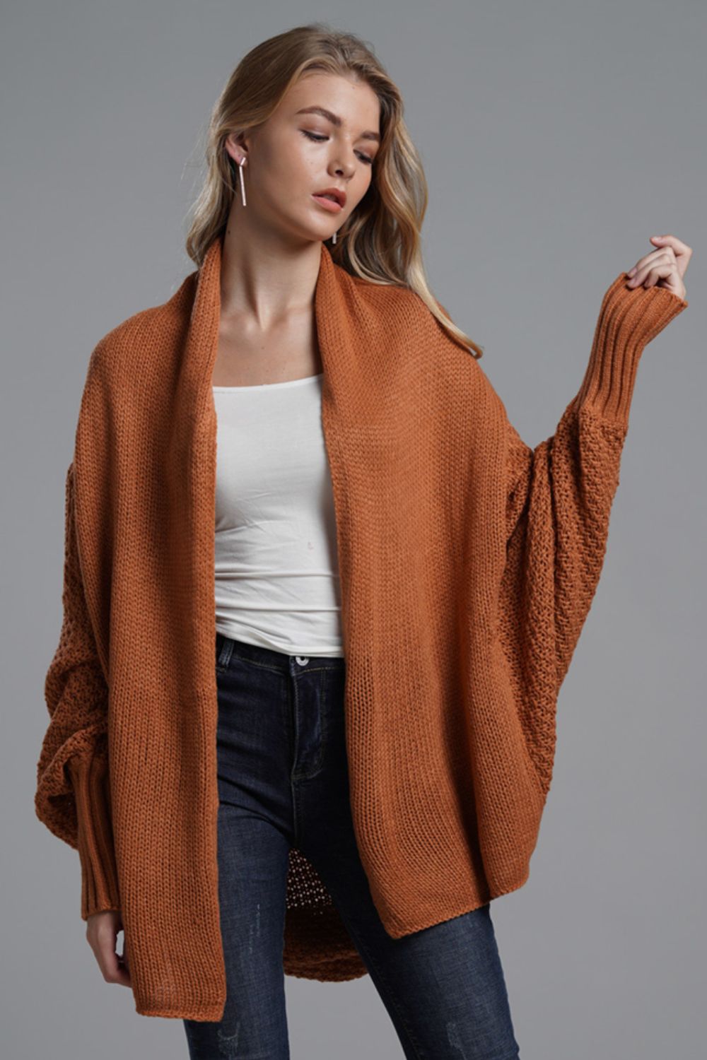 Model wearing a rust-colored dolman sleeve longline cardigan with ribbed details, styled casually with a white top and jeans.