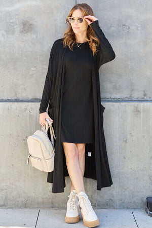 Woman in a black open front long sleeve cover-up, black dress, sunglasses, posing with a backpack.