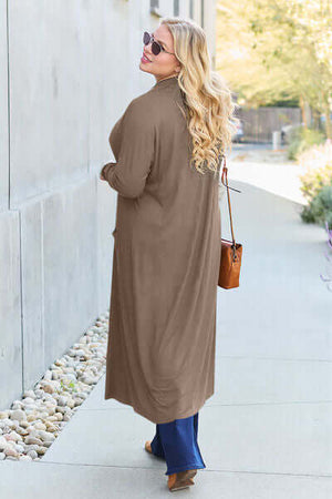 Woman wearing a Basic Bae open front long sleeve cover-up in taupe, walking down a sunny path.