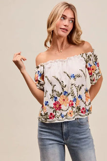 Model wearing a floral embroidered two way off the shoulder blouse with ruffled sleeves and denim jeans.