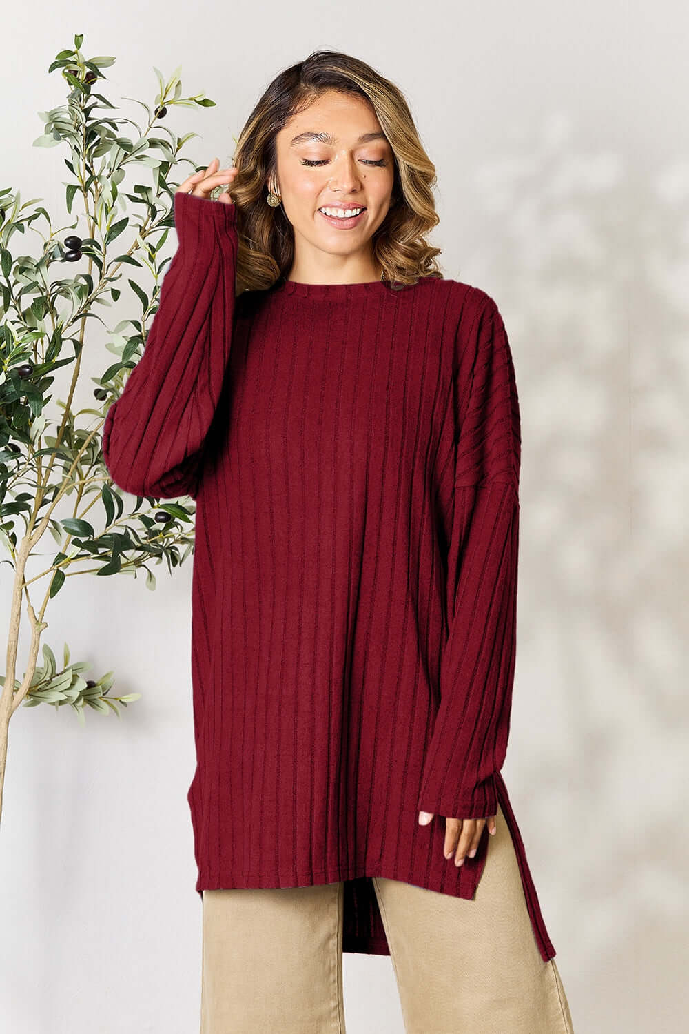 Model wearing a ribbed burgundy long sleeve top with slit, styled with neutral pants and a plant backdrop.