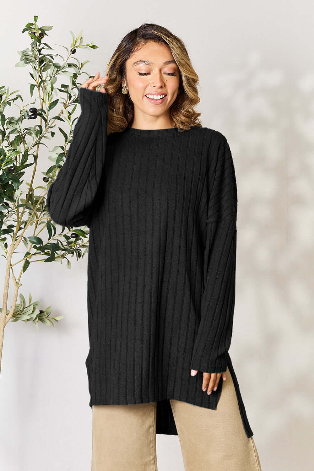 Model wearing a black ribbed long sleeve top with side slits, styled with beige pants and a natural background.