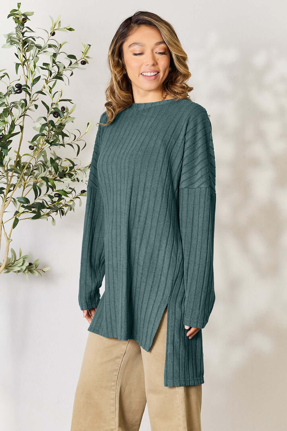 Model wearing a teal ribbed long sleeve slit top, styled with beige pants against a light background.