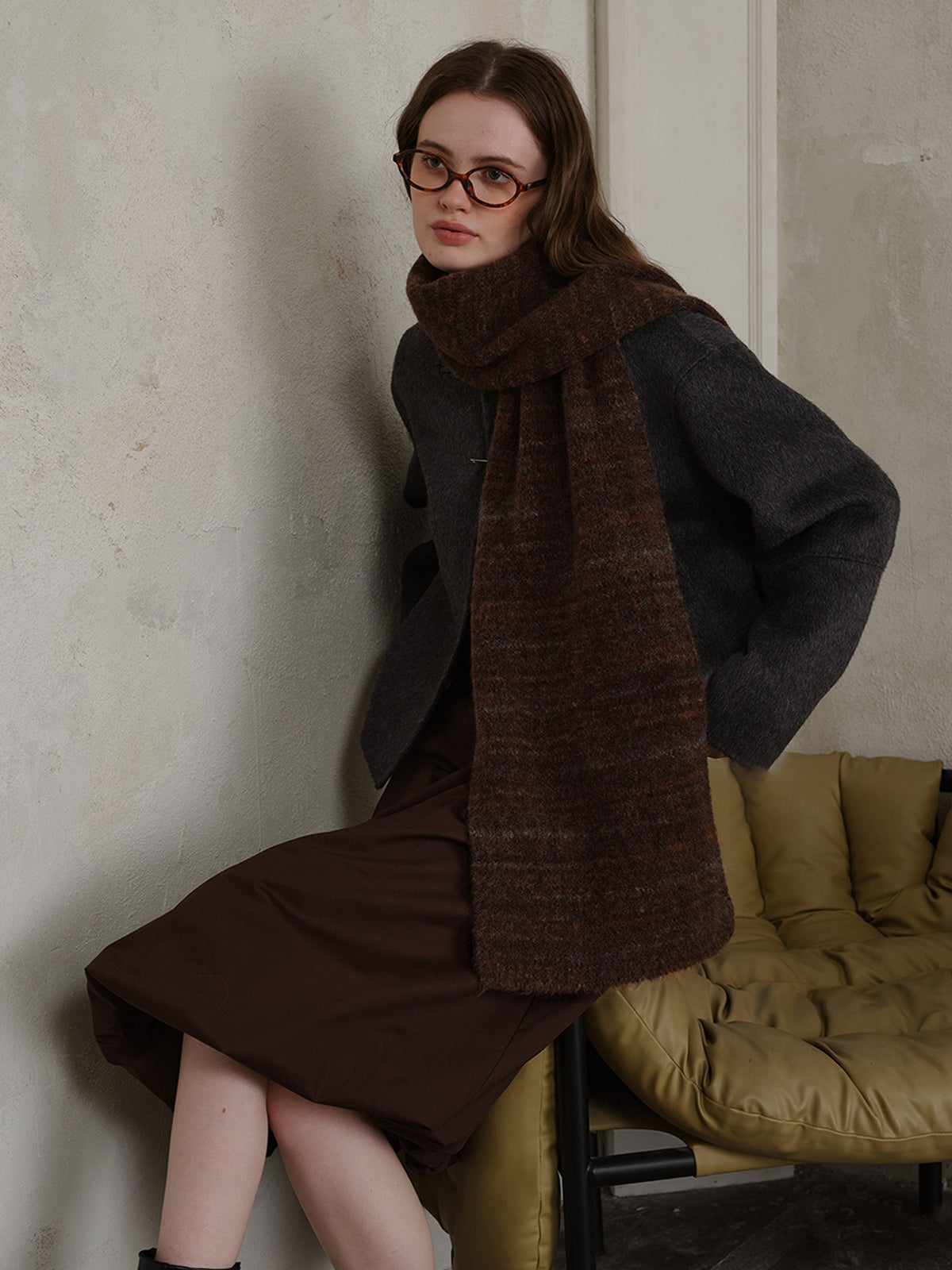 Model wearing a striped detail knit scarf in fur, paired with a stylish gray coat and brown dress, seated elegantly.