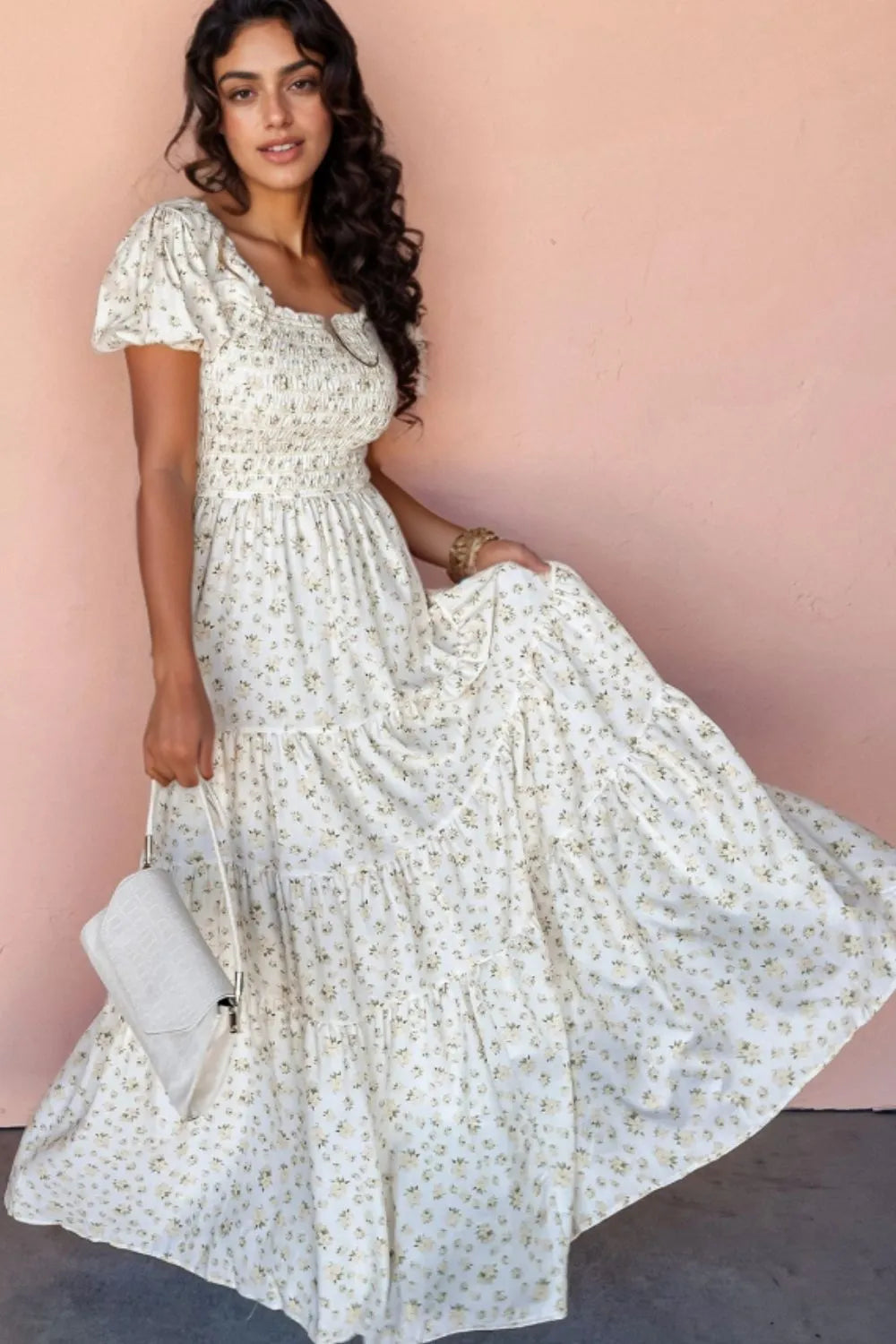 Woman wearing a floral smocked short puff sleeve maxi dress with a tiered design, standing against a pink background.