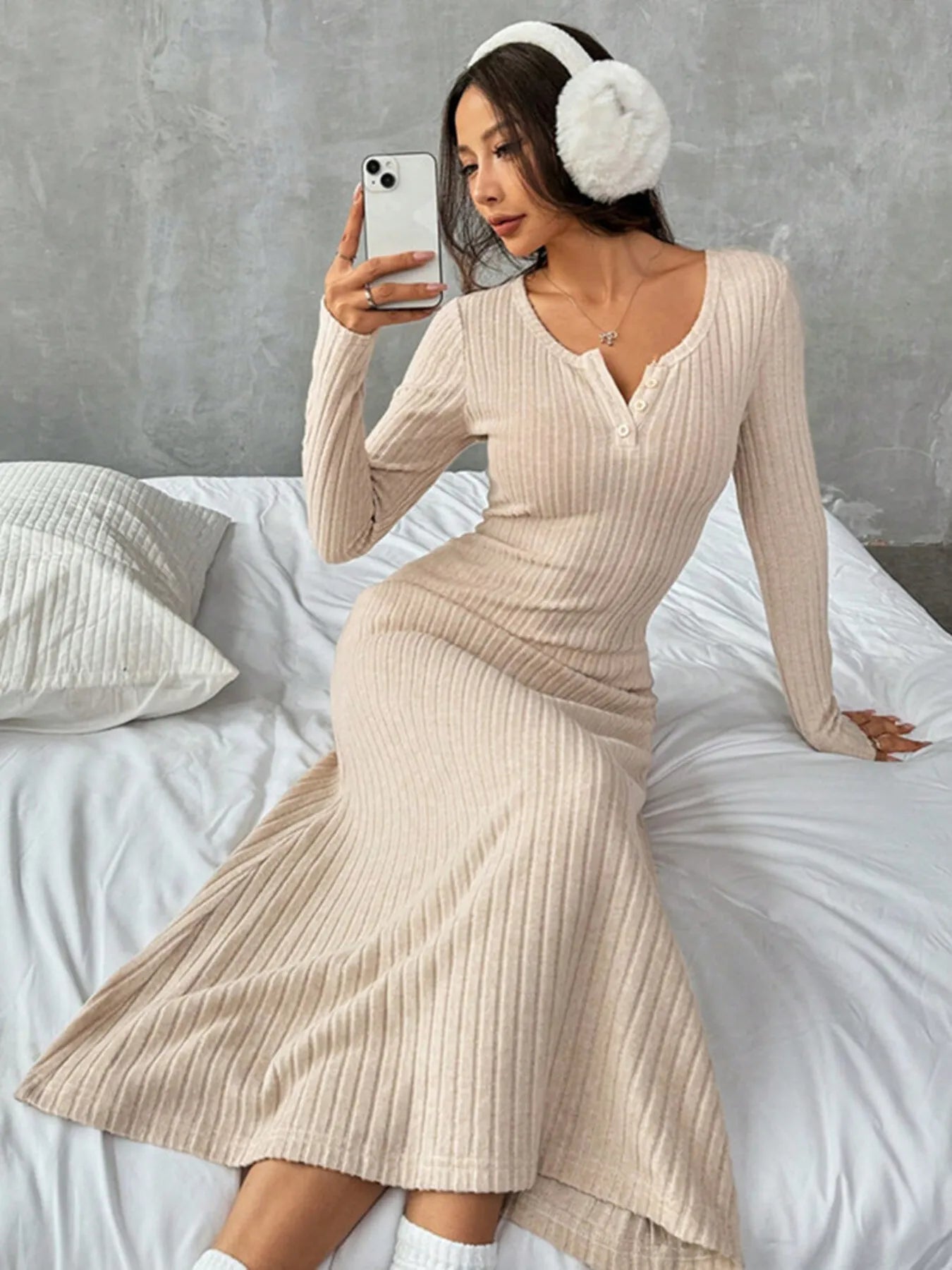 Model wearing a ribbed long sleeve knit dress in beige with button detail, sitting on a bed in a cozy setting.