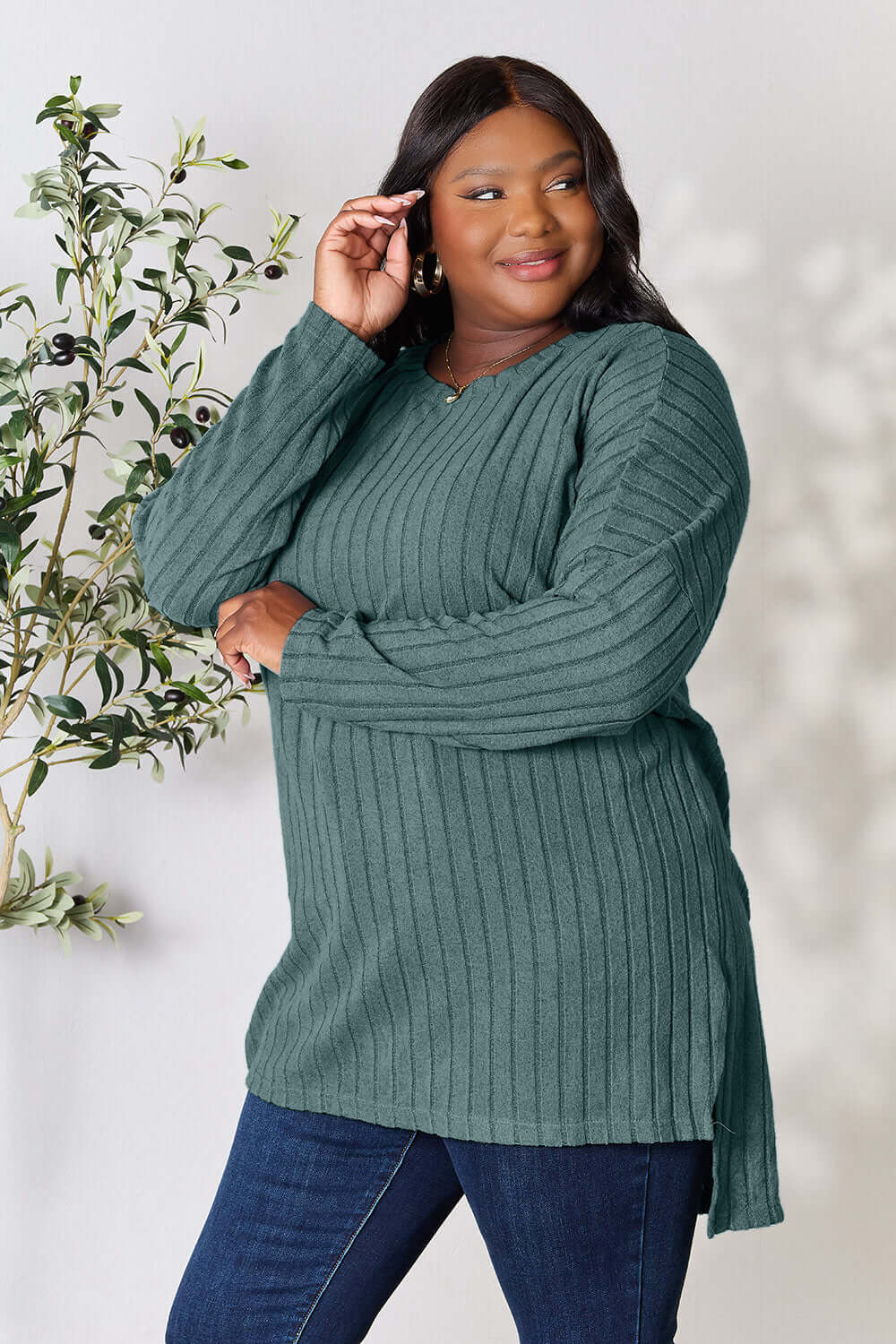 Model wearing a teal ribbed long sleeve slit top with a high-low hem, posing stylishly beside greenery.