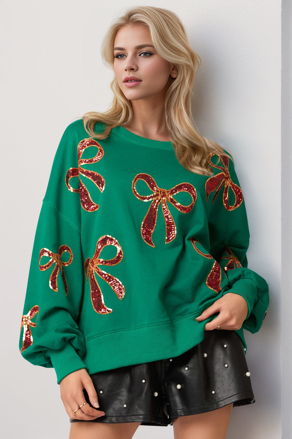 Model wearing a green Double Take Sequin Bow Sweatshirt with red sequin bows and leather shorts.