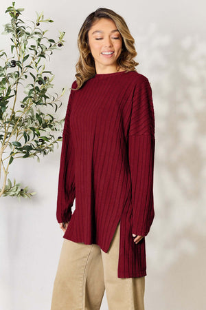 Model wearing a burgundy ribbed long sleeve top with a slit detail, styled with beige pants and a leafy background.