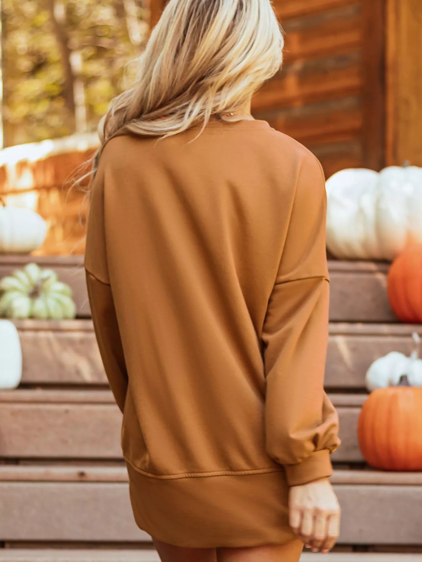 Woman wearing a brown oversized sweater, standing in front of pumpkins and seasonal decor.