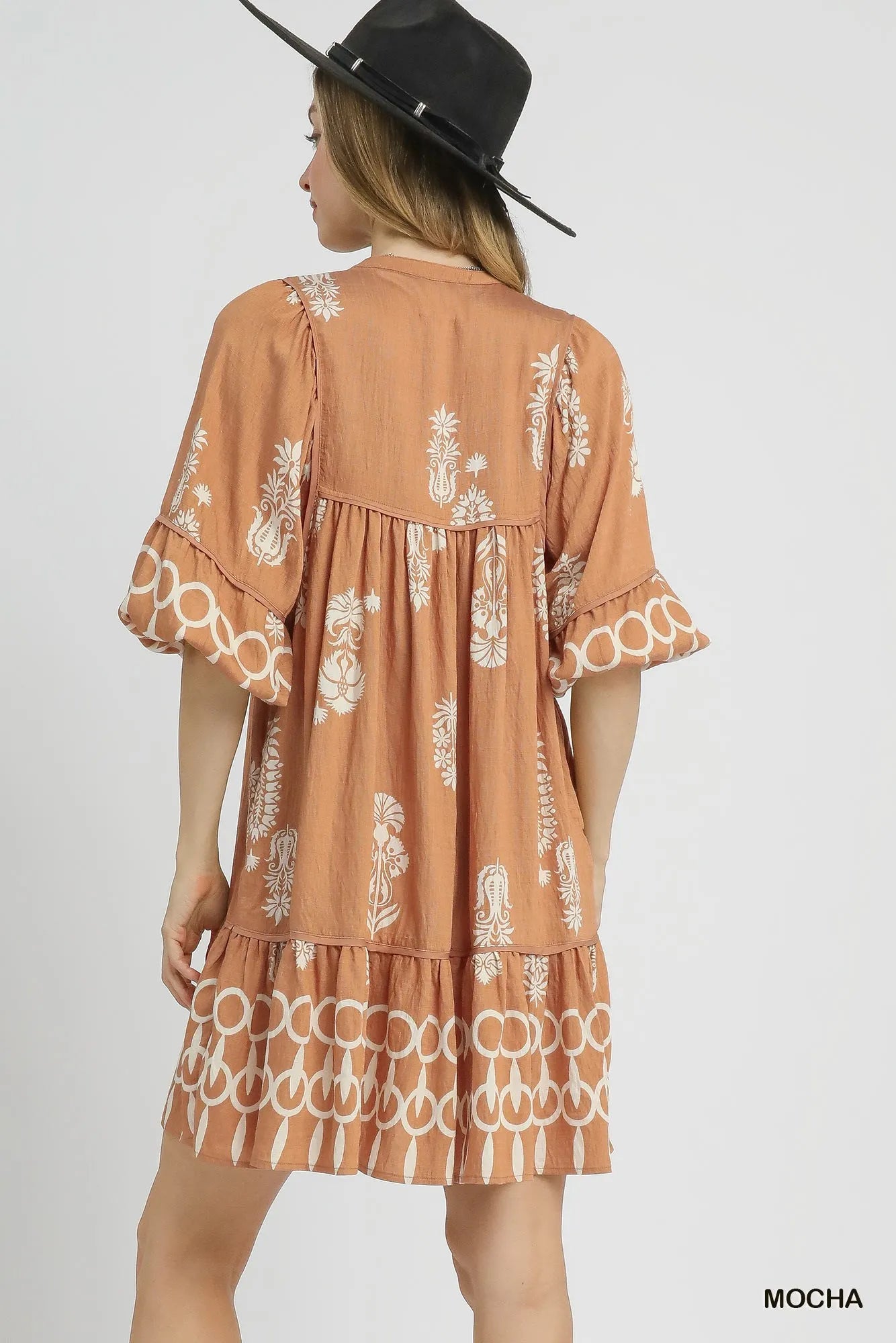 Back view of a woman in a mocha tiered mini dress with ruffled sleeves and floral border print.