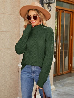 Stylish woman in a green cable-knit mock neck sweater, paired with jeans and a wide-brimmed hat, posing outdoors.