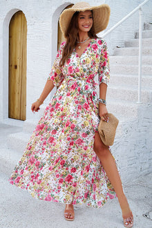 Model in a floral printed tied half sleeve slit dress, wearing a sun hat and holding a bag, standing outdoors.